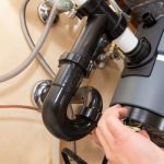 Hand inspecting a garbage disposal unit's plumbing connections, illustrating maintenance and repair services offered by Plumb Perfect Plumbing in Fredericksburg, VA.